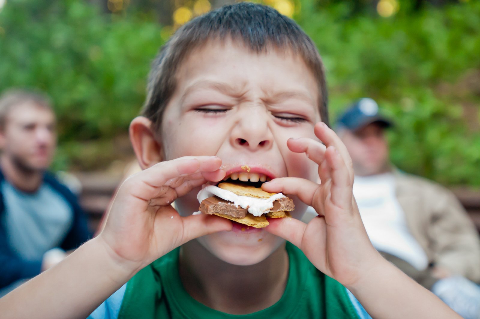 A young boy with short brown hair and a green shirt is eating a s'more outdoors. The boy's eyes are closed, and he appears to be enjoying the treat. In the background, there are two men sitting and a green, leafy outdoor setting.