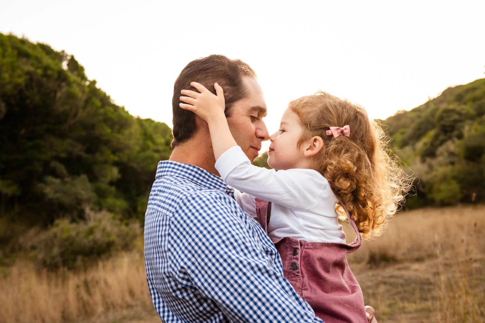 A man holding a young girl close, with their foreheads touching and eyes closed, in an outdoor natural setting during sunset.