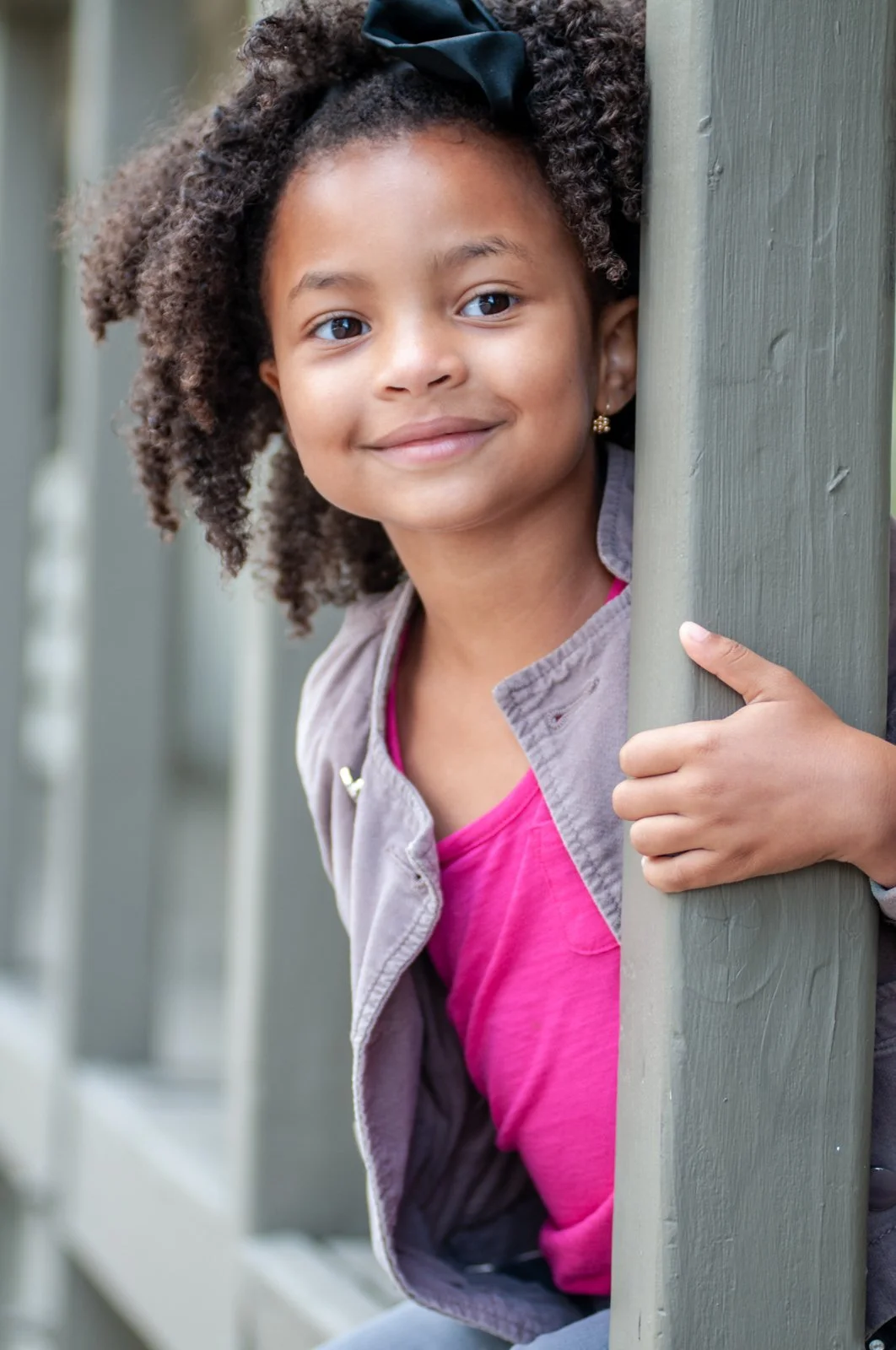 A young girl with curly hair, wearing a dark bow, gray jacket, and pink shirt, peeks out from behind a green wooden post, smiling.