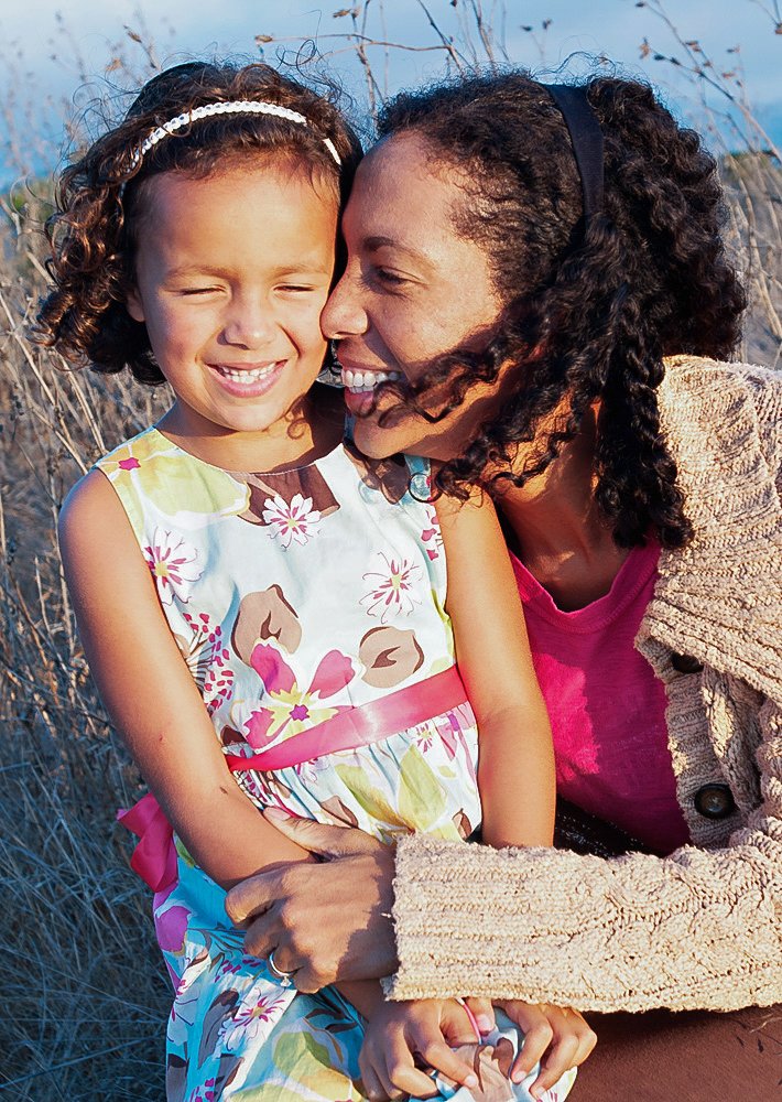 A woman and young girl smiling and hugging outdoors in a grassy field during daytime.