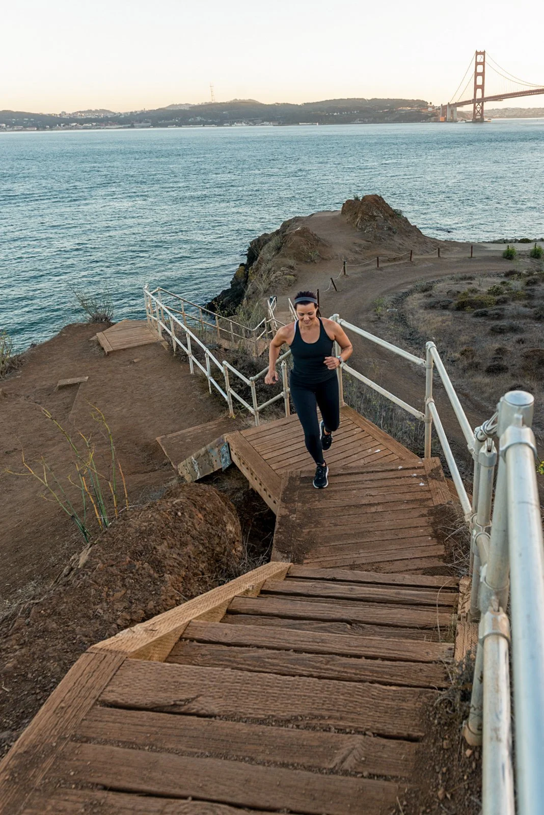 Woman jogging up wooden stairs near the coastline with the Golden Gate Bridge in the background at sunset.