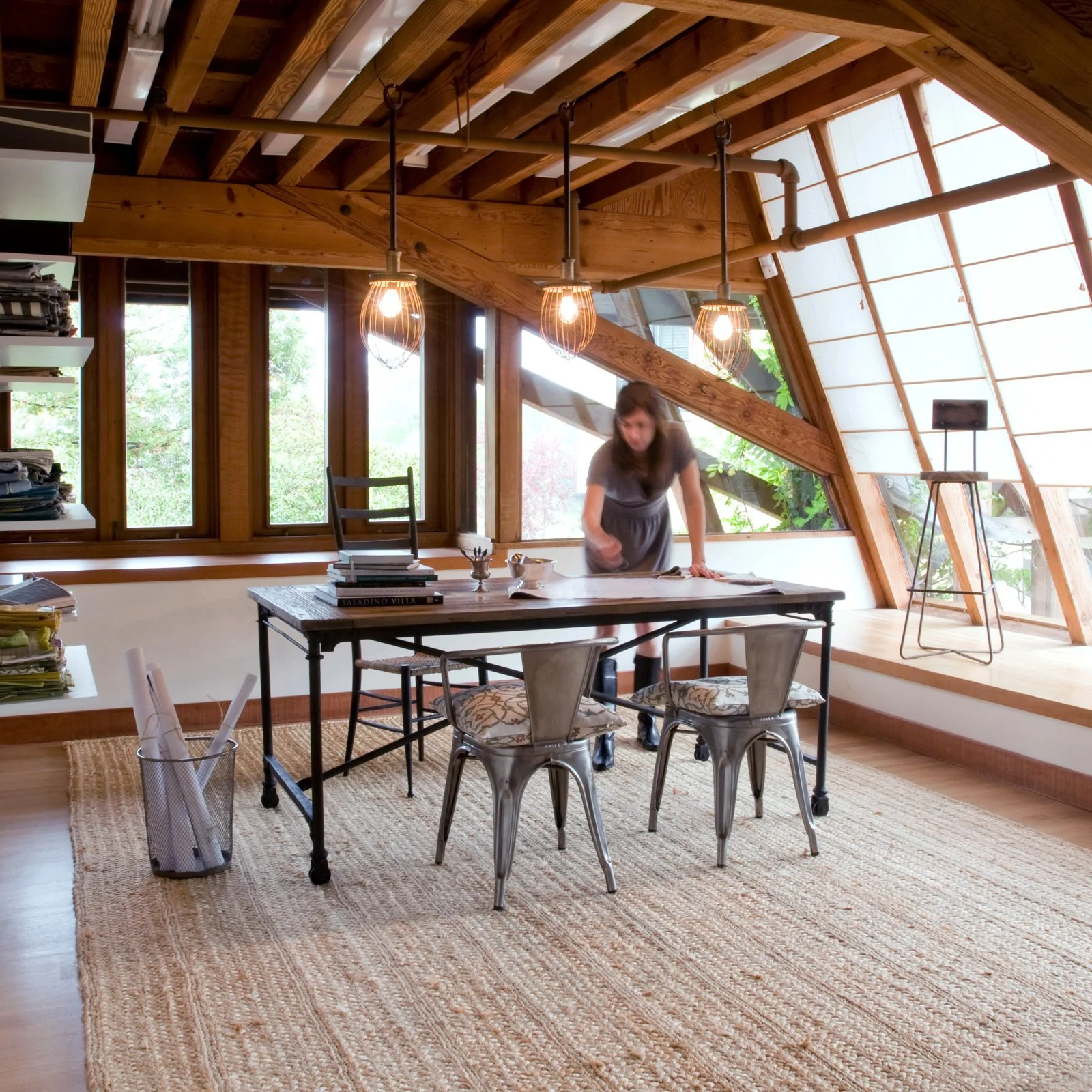 A woman is leaning over a large table in a room with wooden beams, large windows, and modern lighting. There are chairs around the table and a bookshelf with piles of papers on the left side of the image. The room appears bright and airy with natural