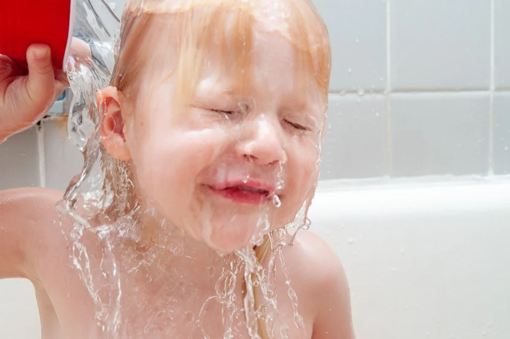 Child with red hair enjoys taking a shower, with water splashing on face and eyes closed.