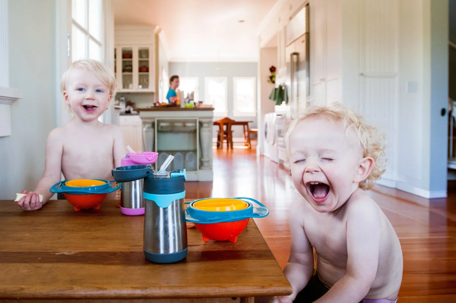 Two young children without shirts are sitting at a wooden table in a bright kitchen. The child on the left is smiling and holding a piece of food, while the child on the right is yawning or making a funny face with eyes closed. There are colorful bow