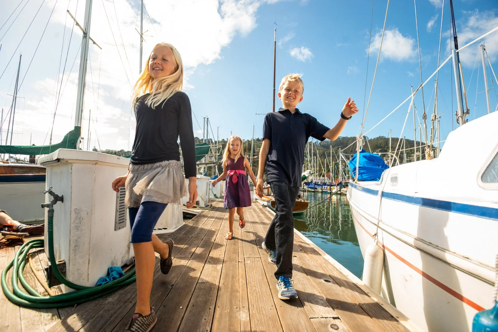 Three children walking and running on a wooden dock at a marina filled with sailboats under a partly cloudy sky.
