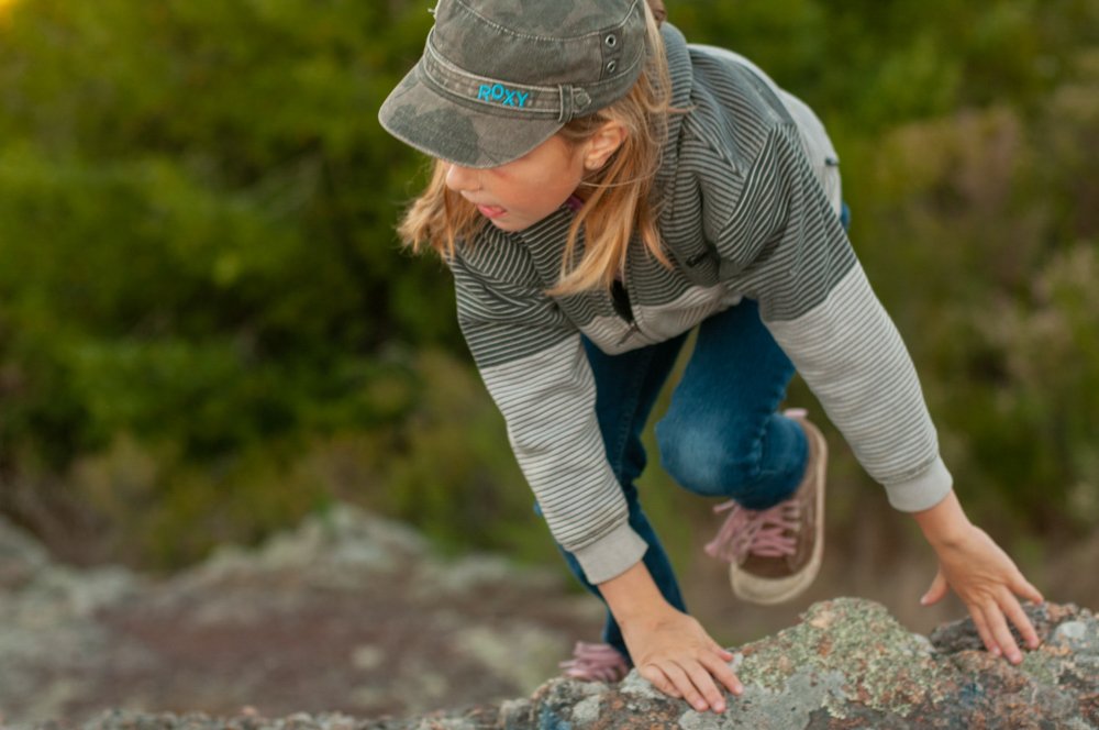 A woman climbing a rocky surface outdoors in a natural setting with green trees in the background.