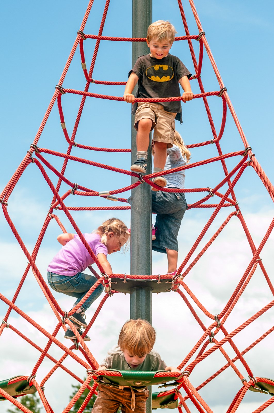 Four children climbing a red rope playground structure on a sunny day.