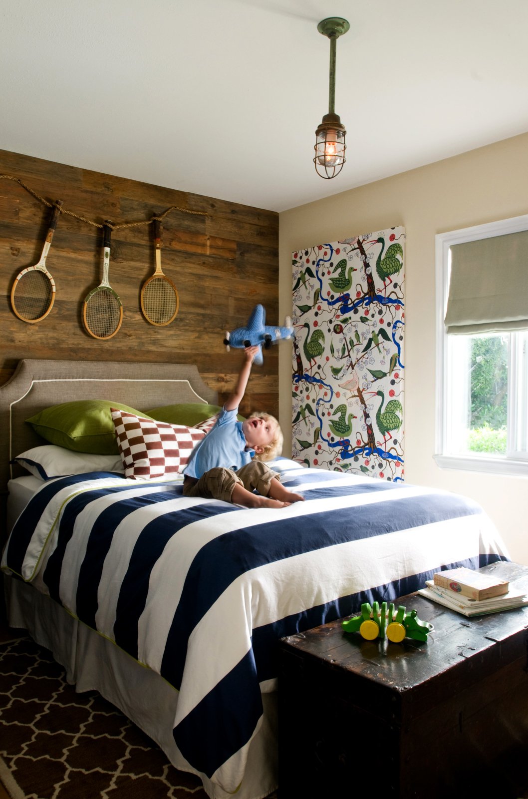 A young boy with blonde hair sitting on a bed with a striped navy and white bedspread, holding a blue plush airplane toy, in a bedroom with a wooden headboard, pillows, a patterned wall hanging, and tennis rackets on the wall.