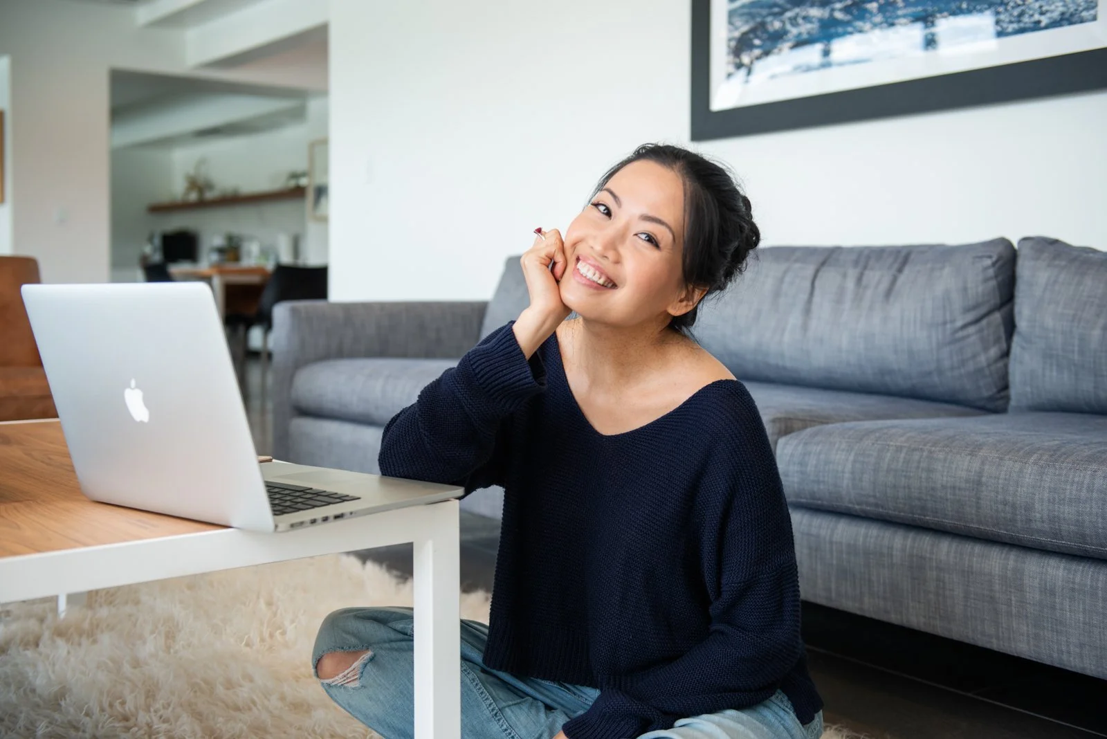 A woman sitting on a fluffy rug in front of a laptop, smiling and resting her chin on her hand inside a living room.