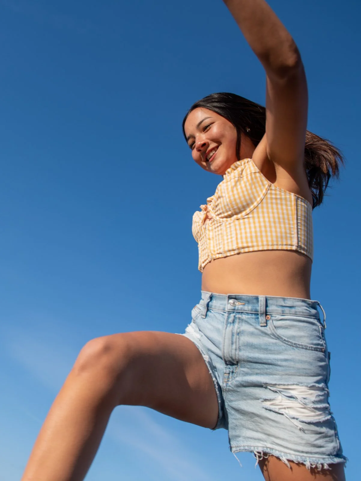 A young woman smiling outdoors against a clear blue sky, wearing a yellow checkered crop top and ripped denim shorts.