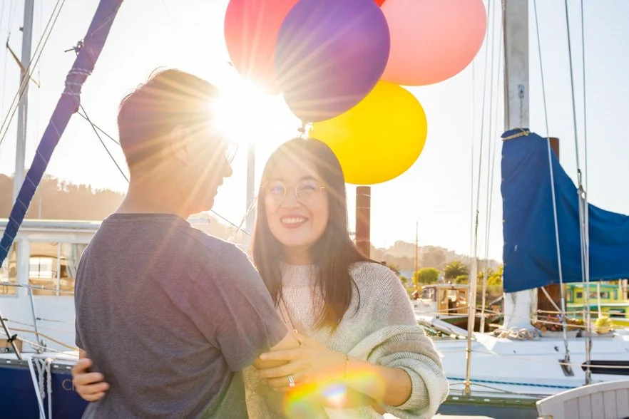 Two people smiling and holding balloons on a boat dock, with sailboats and a setting sun in the background.