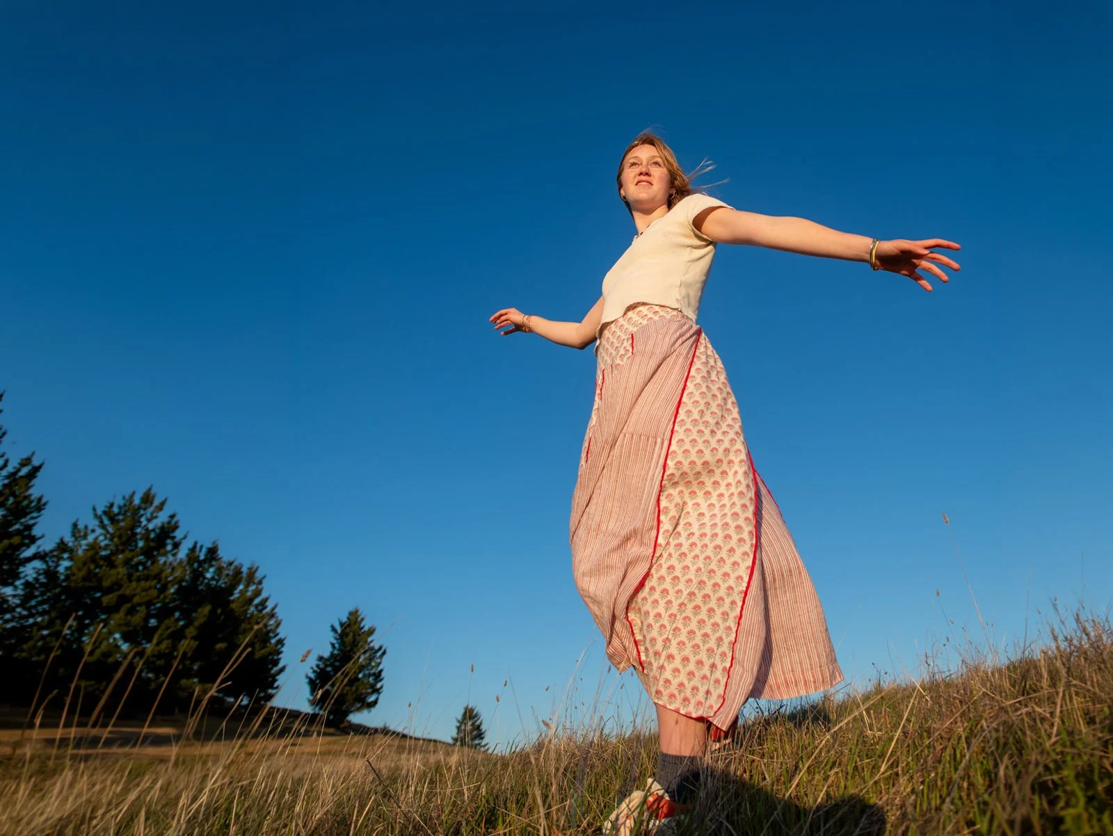A woman standing outdoors on a grassy hill, wearing a beige top and a long patterned skirt, with her arms outstretched, looking up at a clear blue sky.