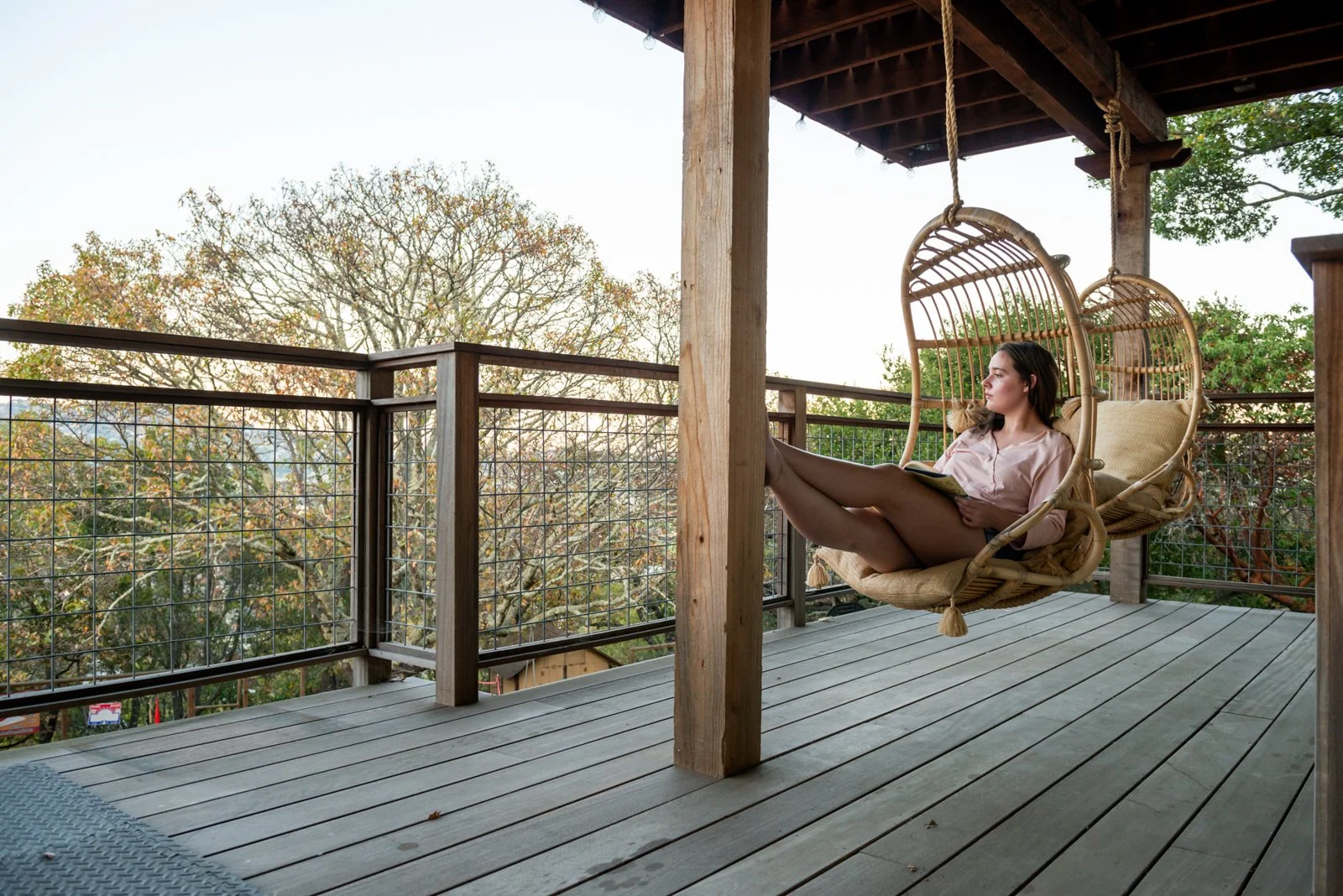 A woman sitting in a hanging wicker chair on a wooden balcony, reading a book, with trees and a clear sky in the background.