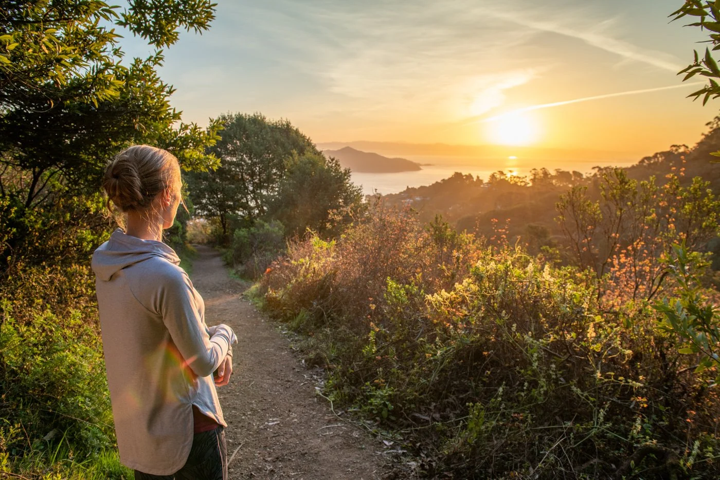 Woman standing on a dirt trail looking at a sunset over a landscape with trees and a body of water in the distance.