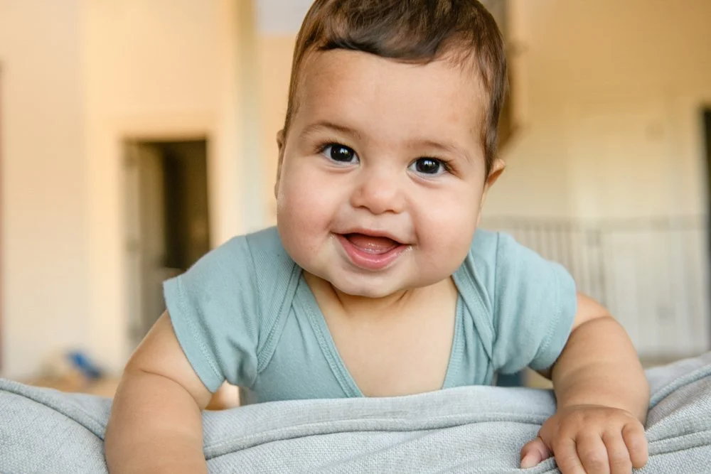A smiling baby with dark hair and big eyes, wearing a light blue shirt, leaning on a soft surface indoors.