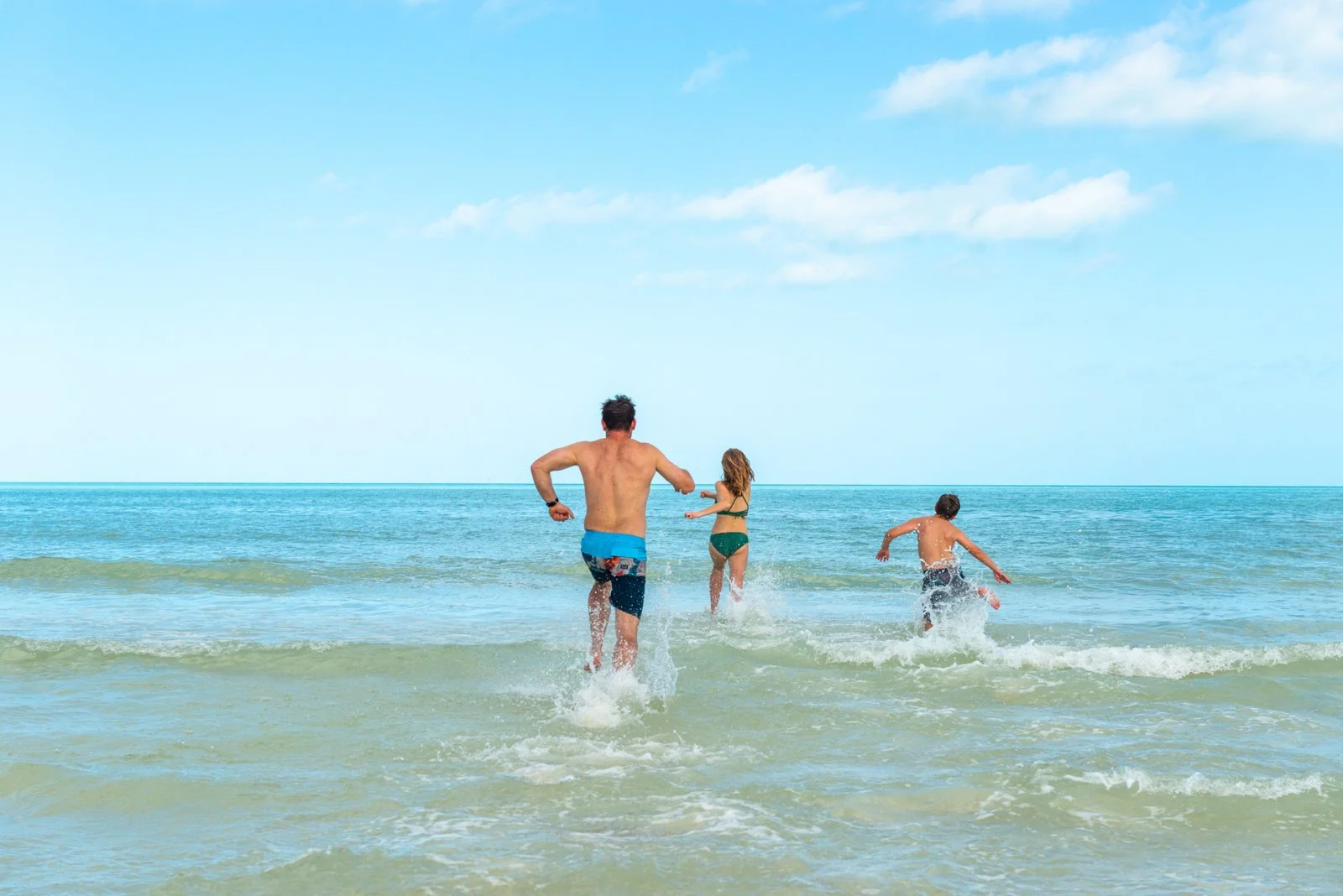Family of three playing in the ocean on a sunny day, with blue sky and clouds, in swimwear.