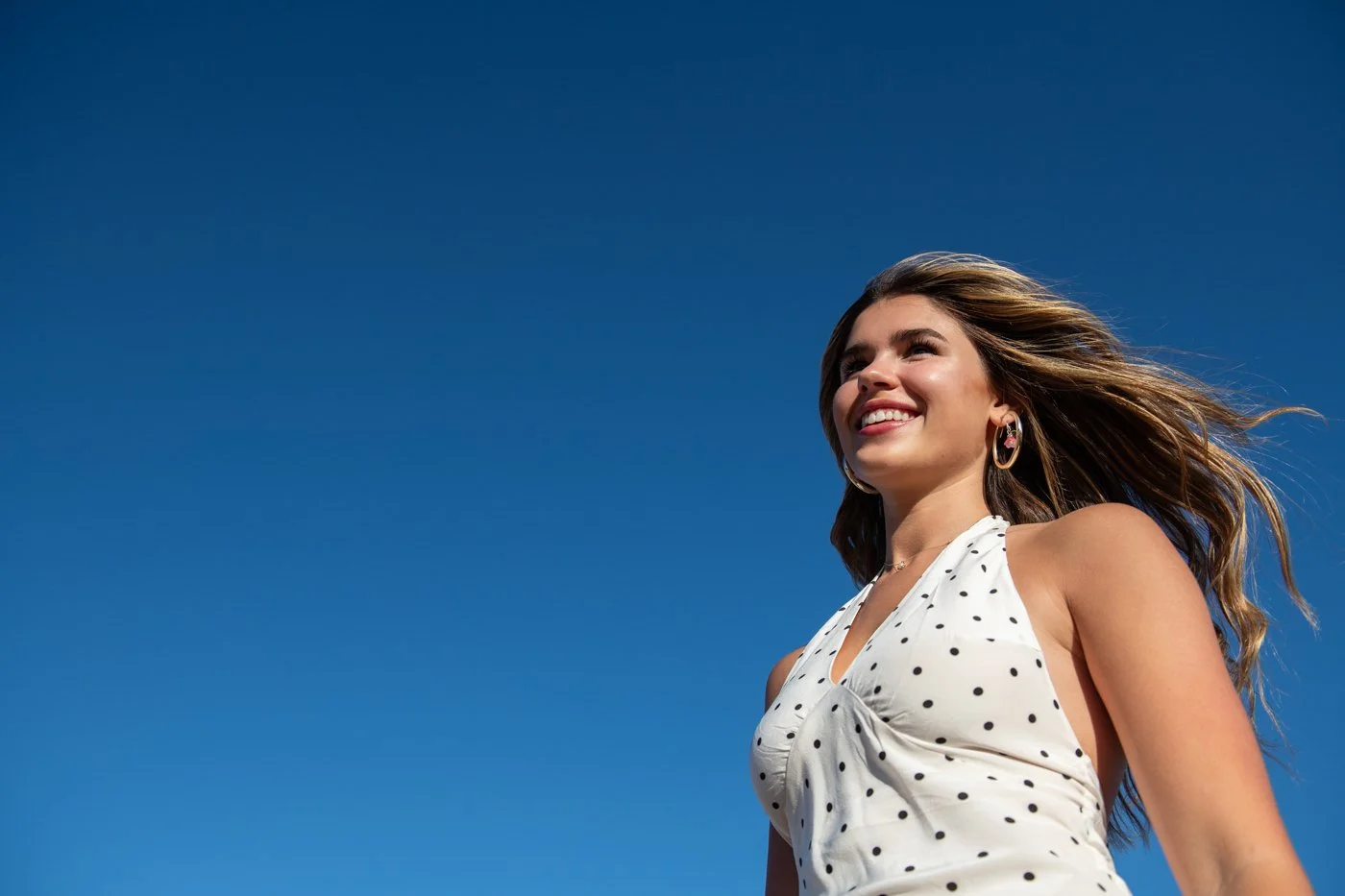 A young woman with long wavy hair, smiling, looking up, wearing a white sleeveless dress with black polka dots, earrings, against a clear blue sky.