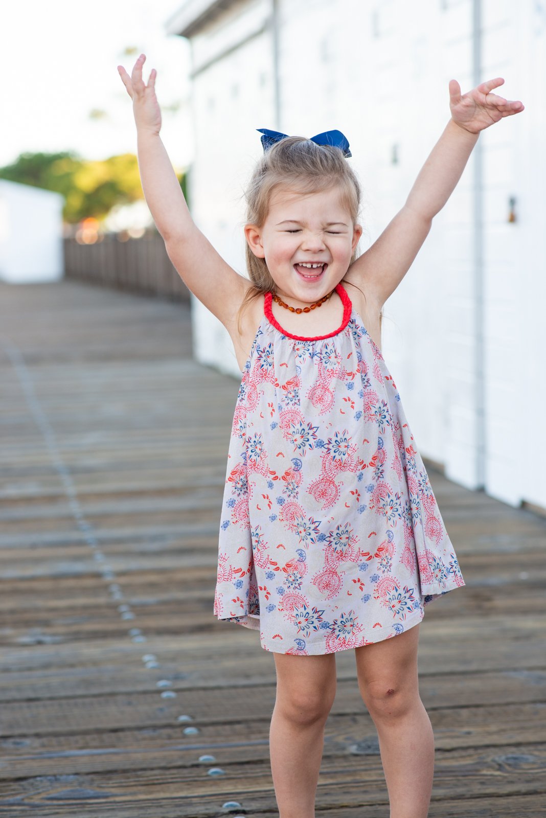 A young girl in a floral dress with a blue bow in her hair, smiling with her arms raised up.