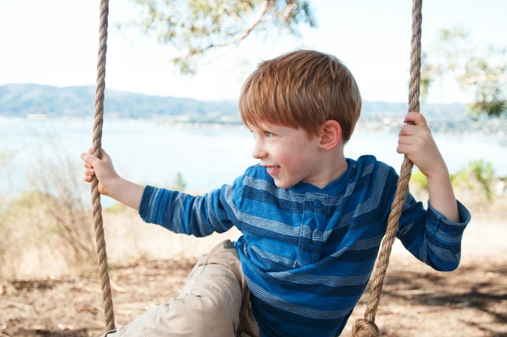 A young boy with red hair, wearing a blue striped shirt, is sitting on a swing outside near water and trees, smiling as he looks to the side.