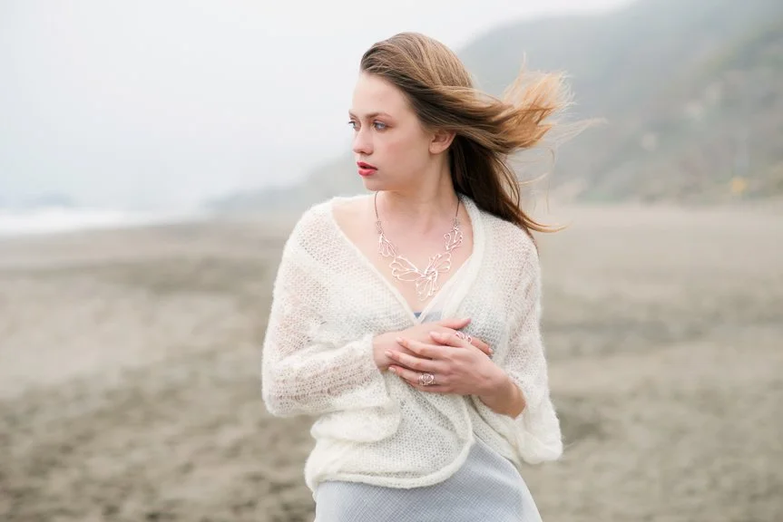 A woman standing on a beach with her hair blowing in the wind, wearing a cream-colored cardigan and jewelry.
