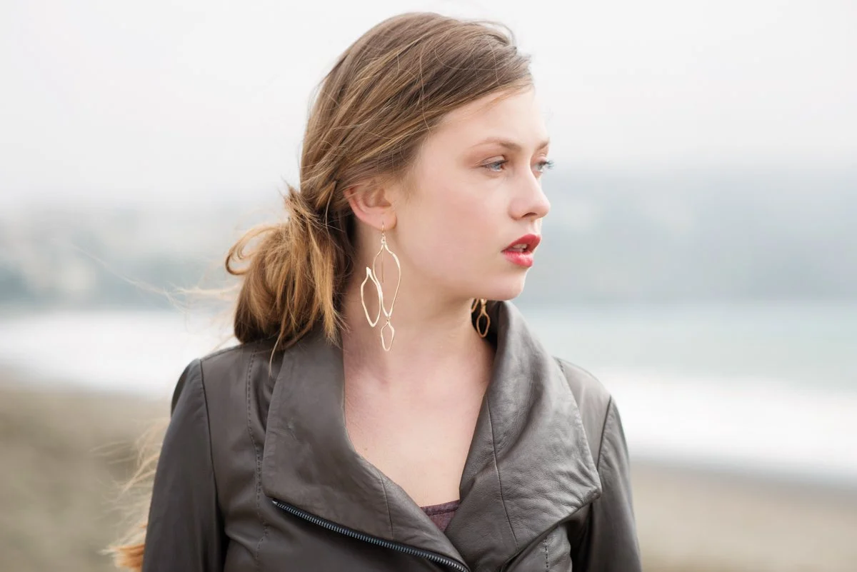 A young woman with light brown hair wearing large gold earrings and a gray leather jacket, standing outdoors near the beach.