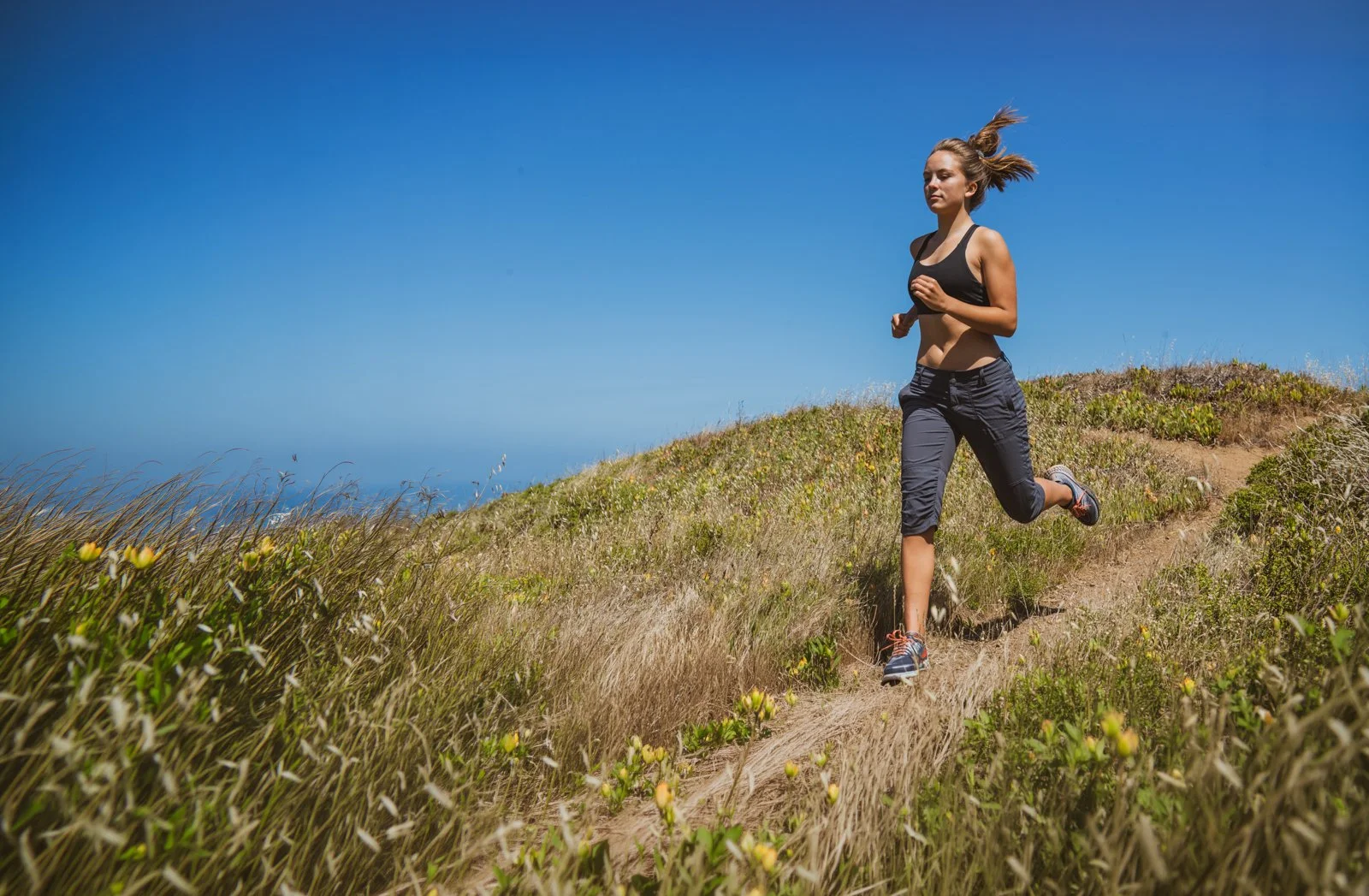 A woman running on a narrow dirt trail through grassy hills with a clear blue sky overhead.