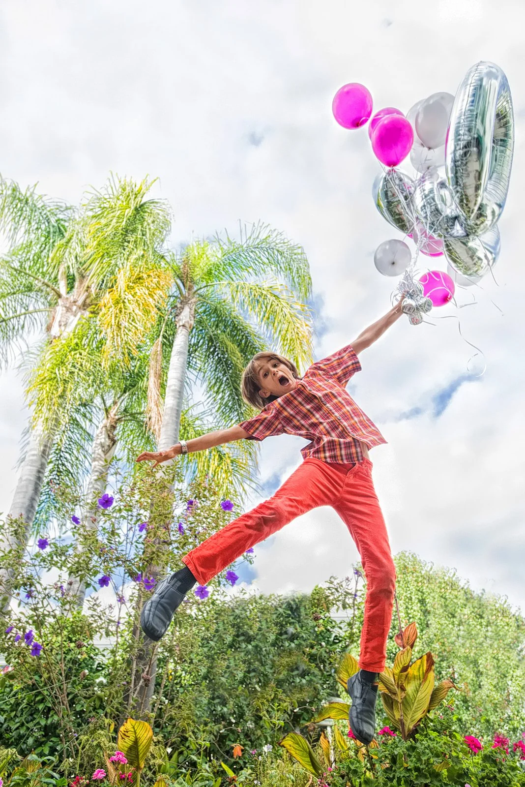 Young boy jumping in a garden holding pink, white, and silver balloons, with palm trees and colorful flowers in the background.