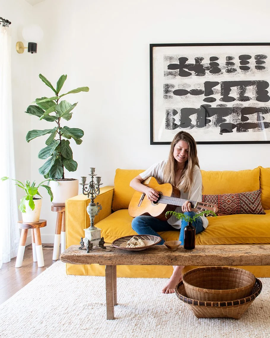 A woman sitting on a yellow sofa playing an acoustic guitar in a living room with a large potted plant, a framed abstract art piece, a wooden coffee table with a tray of snacks, a glass bottle with greenery, and a large empty basket on the floor.