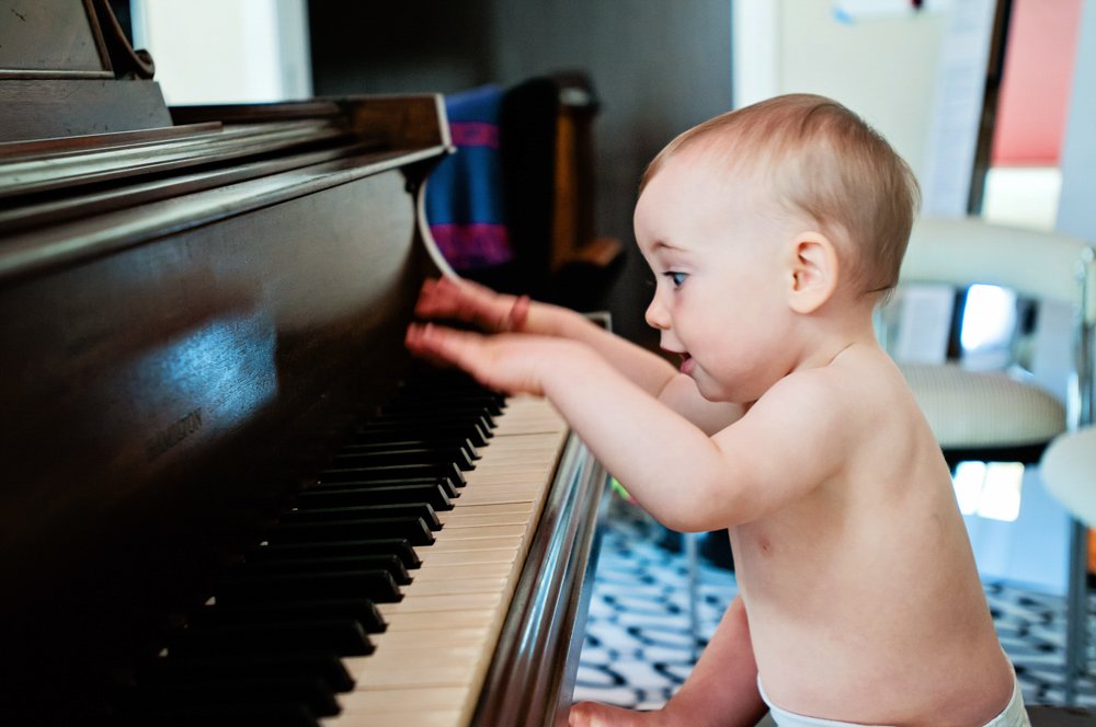 A young baby with no shirt playing a piano, with a focused expression, in a home setting.