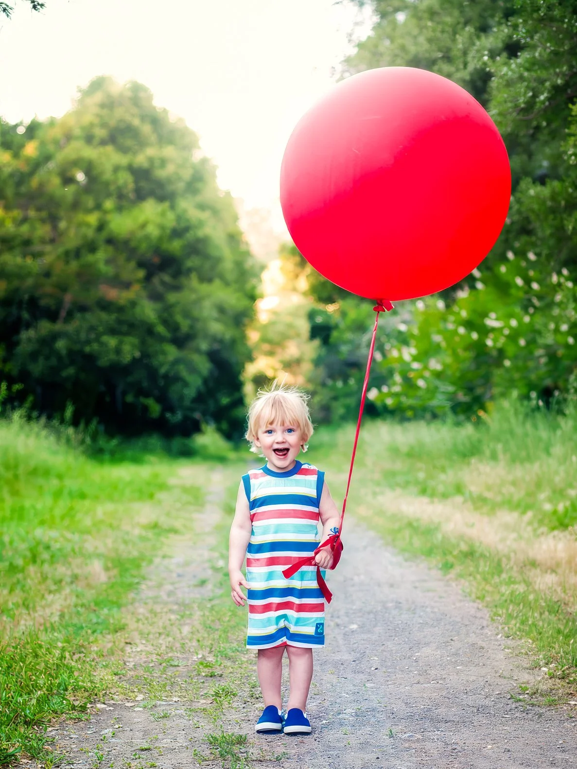 A young boy in a striped shirt and shorts holding a large red balloon on a string, standing on a grassy and dirt path in a green outdoor setting, with trees in the background.