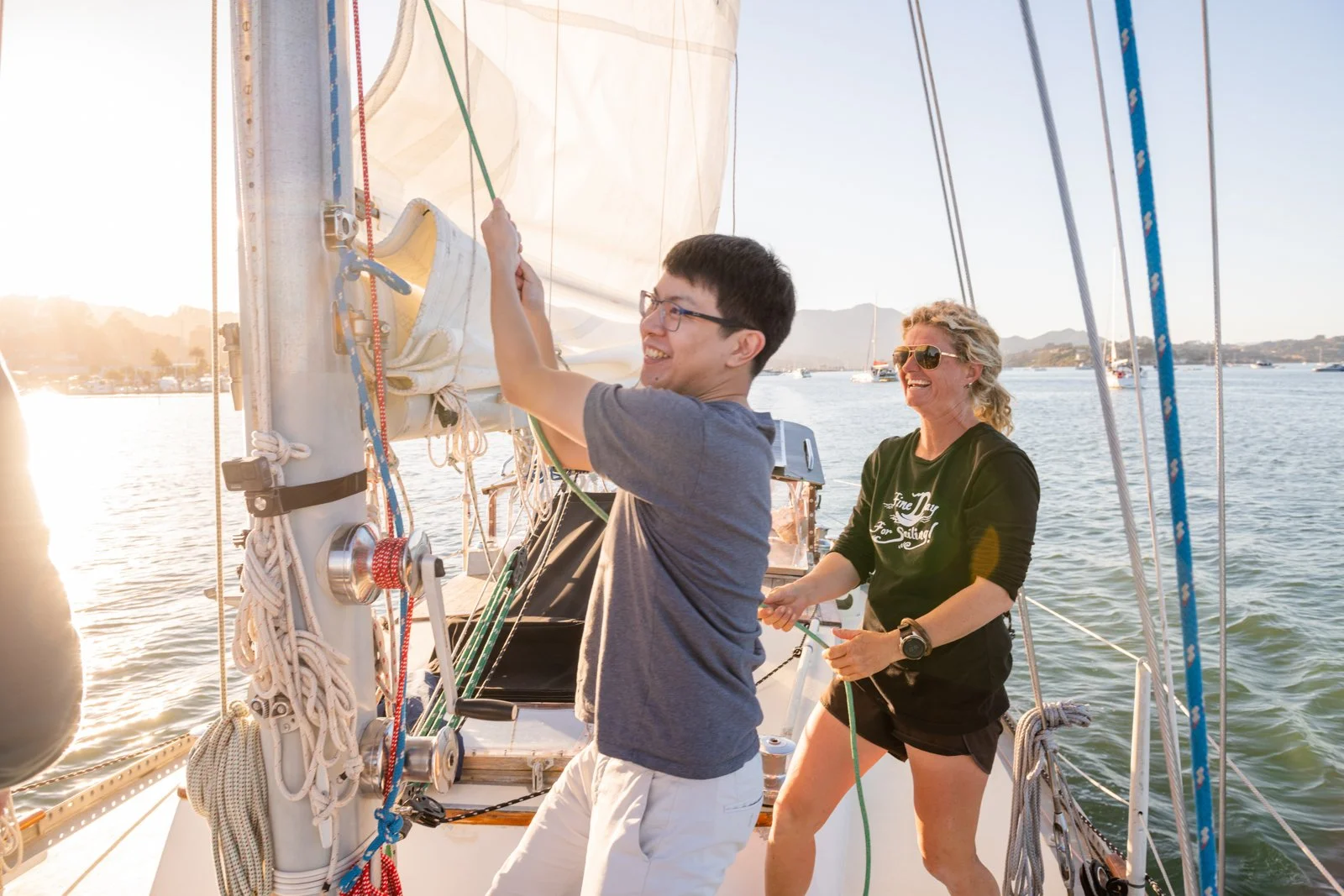 Two people on a sailboat smiling and enjoying the sunset. The person in the foreground is a man with glasses, a blue t-shirt, and light-colored shorts. The woman behind him is wearing sunglasses, a black long-sleeve shirt, and shorts, holding a rope.