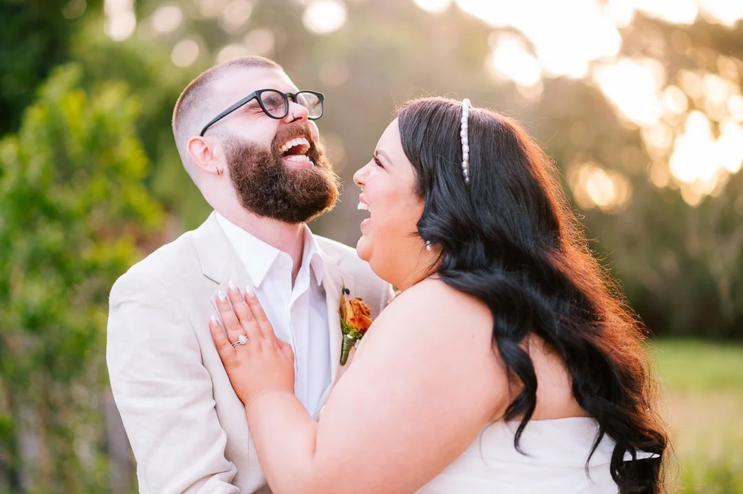 A happy couple, man with glasses and beard and woman with long black hair and headband, laughing and embracing outdoors during sunset.