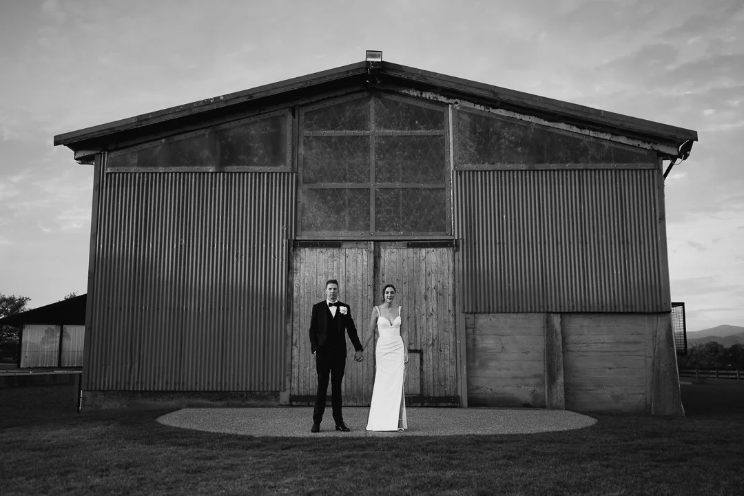 A black and white photo of a bride and groom standing hand in hand in front of a rustic barn.