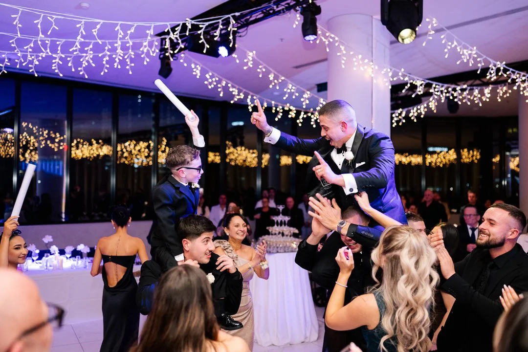 People celebrating at a wedding reception, some lifting a man in a tuxedo, with string lights hanging above.