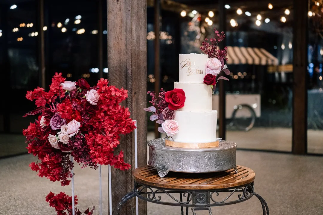 A three-tiered white wedding cake decorated with red and pink flowers, placed on a decorative silver cake stand. To the left, there is a large floral arrangement with red, pink, and purple flowers. The setting appears to be a rustic indoor venue with