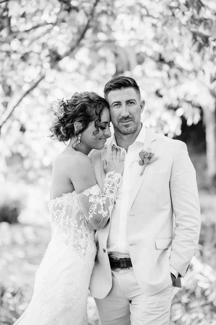 A black and white photograph of a bride and groom standing close together outdoors. The bride is wearing a lace off-the-shoulder wedding dress with floral details, her eyes closed and head resting against the groom's. The groom is dressed in a light