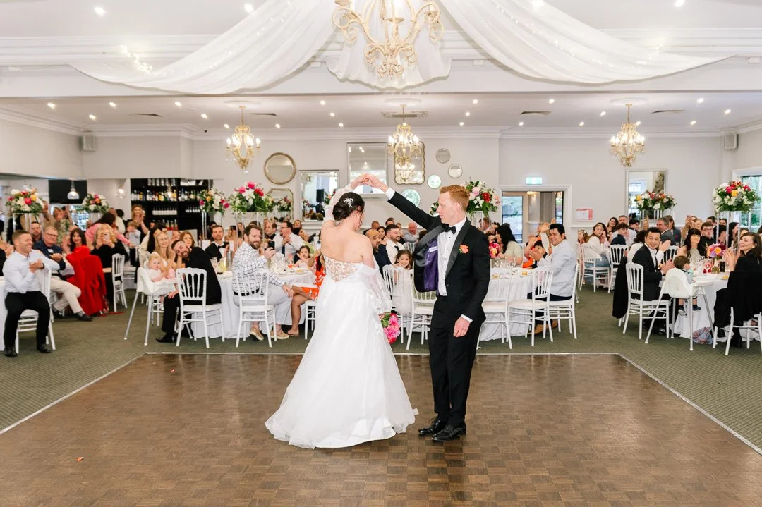 A bride and groom dancing on a wedding reception dance floor, surrounded by seated guests at decorated tables with floral centerpieces in a banquet hall.
