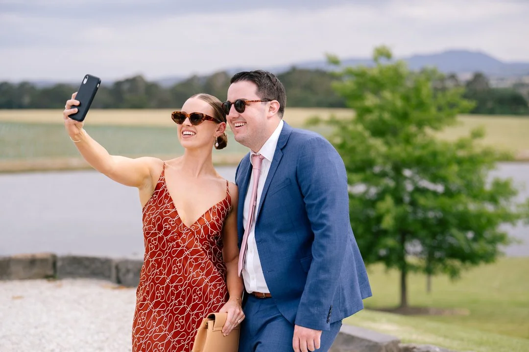 A woman and man dressed in business attire taking a selfie outdoors near a lake, both wearing sunglasses, with green trees and hills in the background.