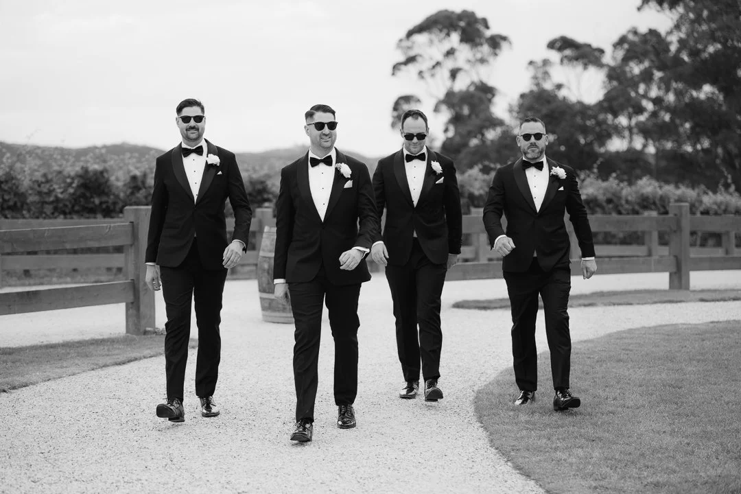 Four men dressed in tuxedos and sunglasses walking on a gravel path outdoors, with trees in the background, during daytime.