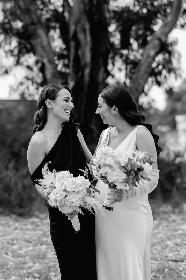 Two women in wedding attire smiling and holding bouquets, outdoors in front of a large tree.