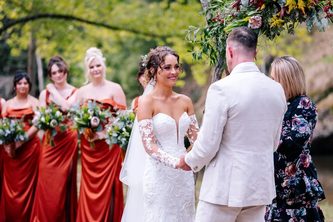 Bride and groom exchanging vows outdoors at wedding ceremony, with bridesmaids in orange dresses holding bouquets in the background.