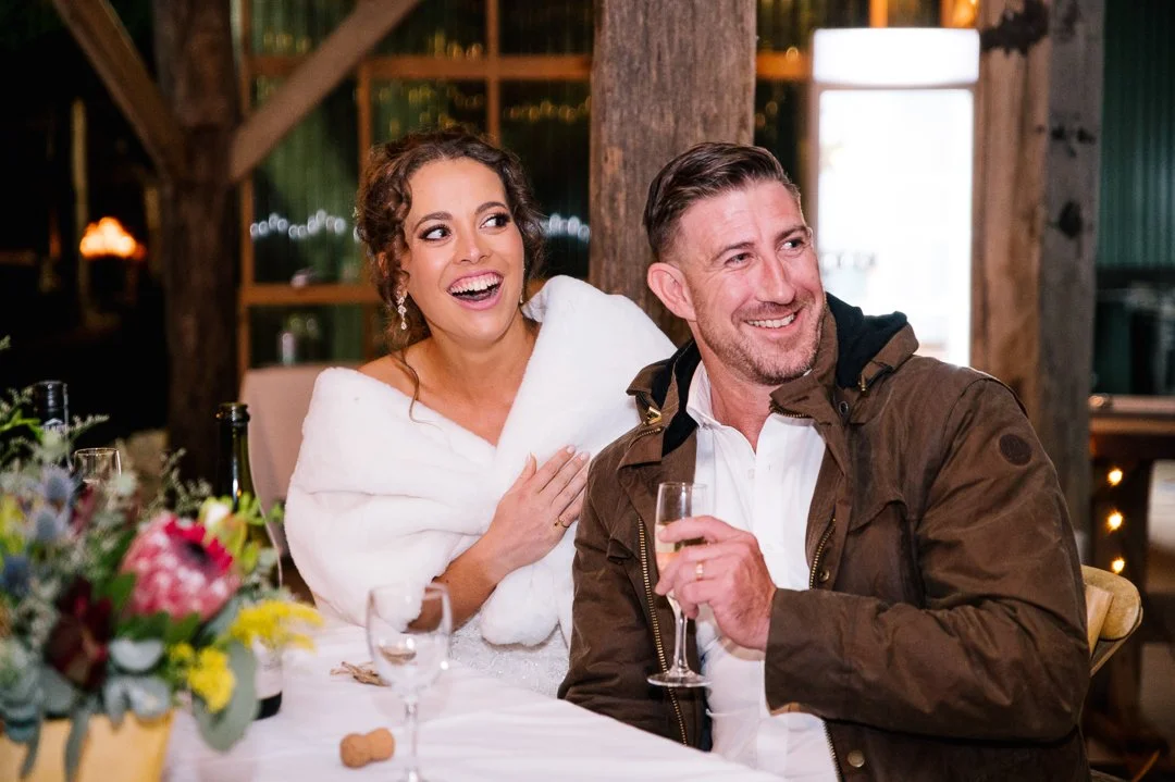 A woman in a white dress and a woman in a brown jacket smiling and laughing at a table decorated with flowers, with glasses of drinks, in a rustic indoor setting.