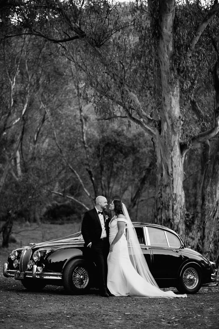 Black and white photo of a bride and groom standing close together next to a vintage black car in a wooded outdoor setting.