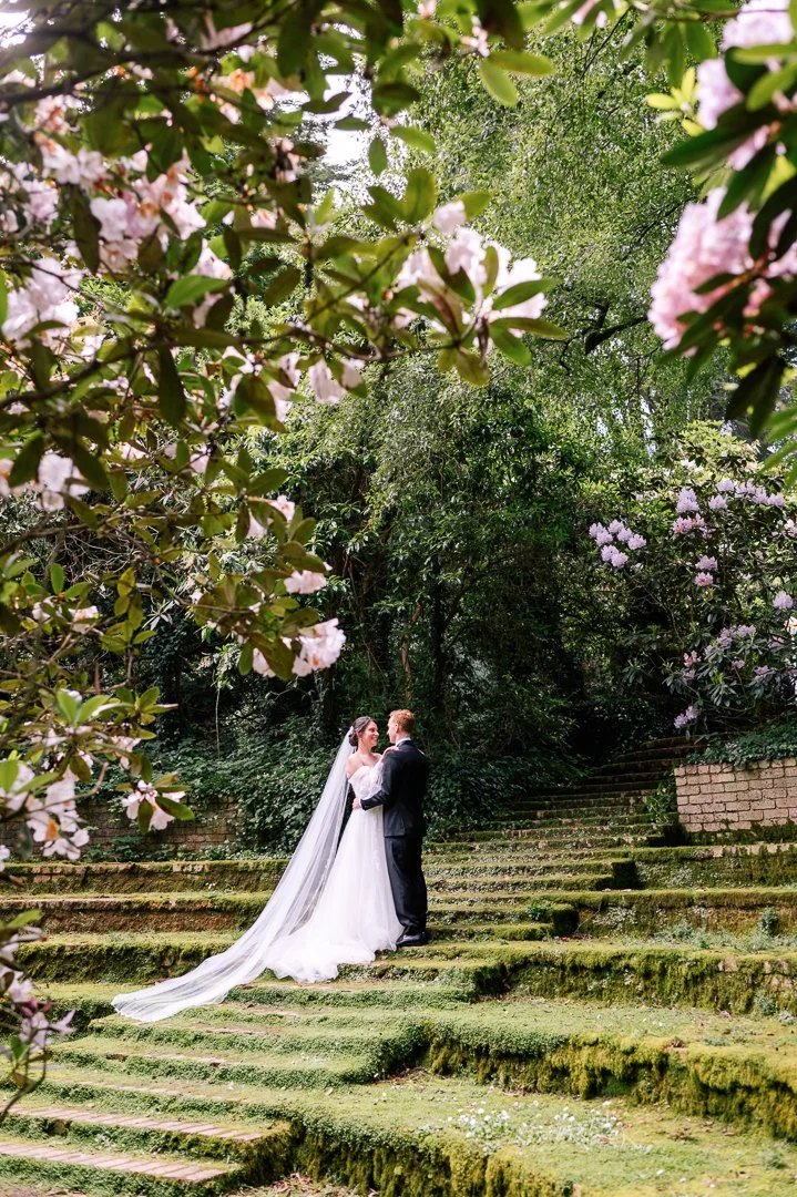 Bride and groom standing on moss-covered stone steps in a lush garden, surrounded by trees and pink blooming flowers, sharing a wedding moment.