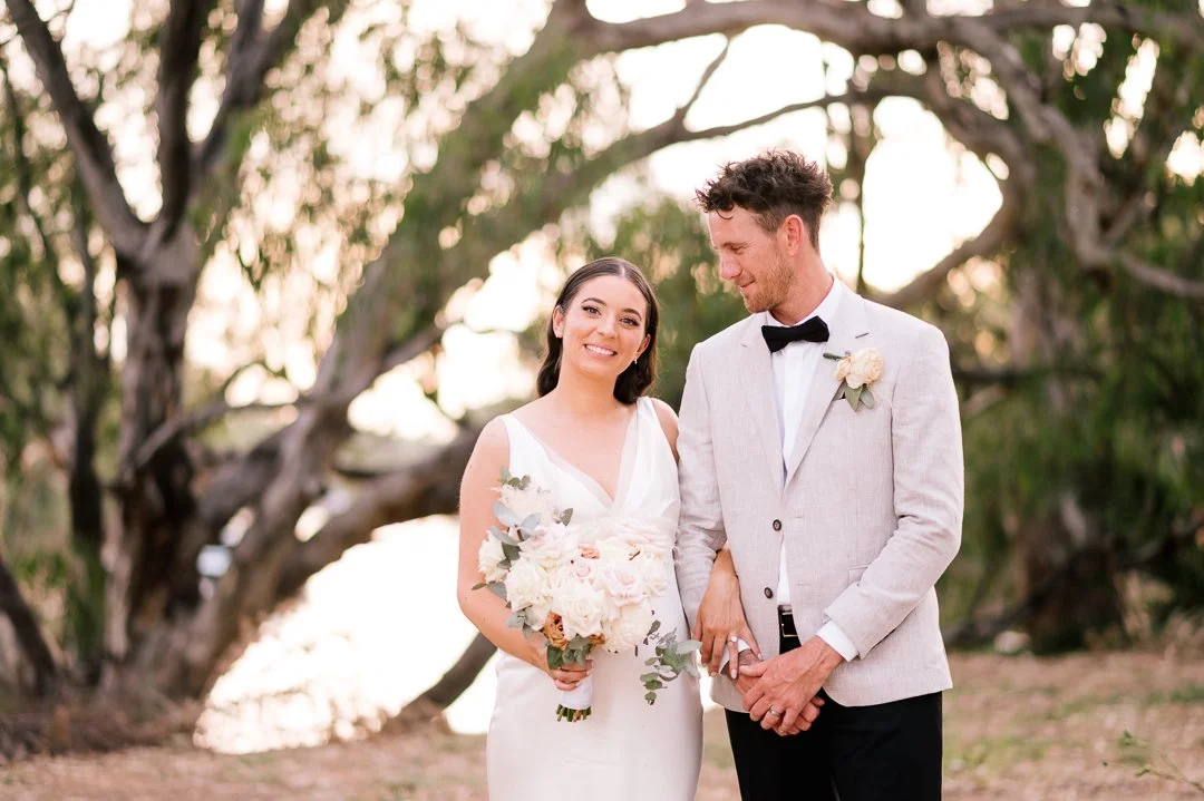 A bride and groom standing outdoors among trees during sunset, with the bride holding a bouquet and both smiling.