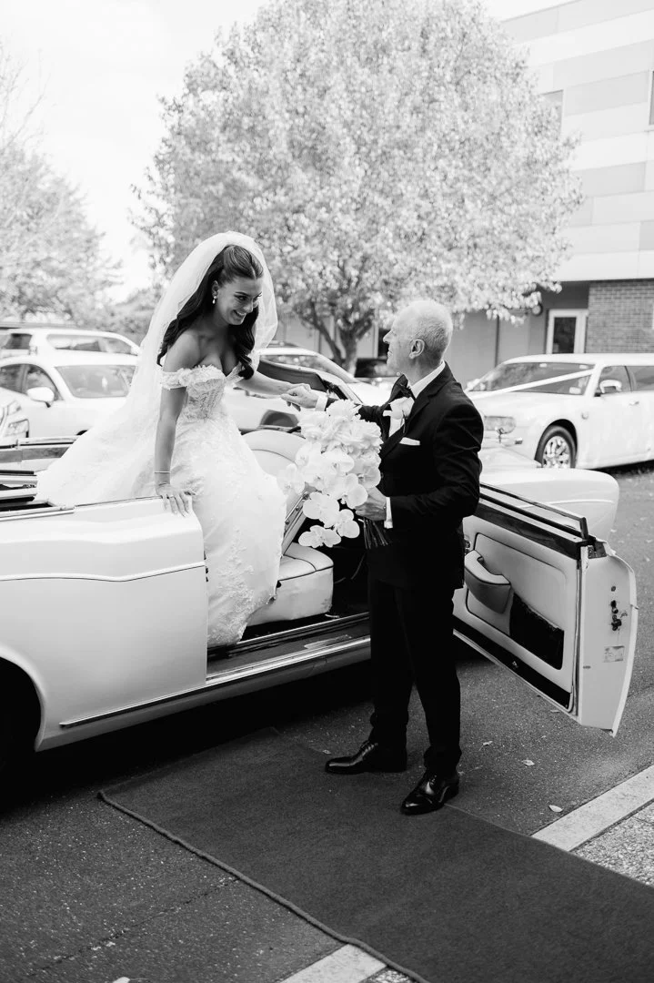 Bride in a wedding dress stepping out of a vintage car, receiving flowers from a man in a tuxedo, outdoors near parked cars and trees.