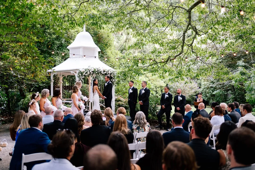 Outdoor wedding ceremony under a leafy tree, with a white gazebo decorated with flowers. Bride and groom hold hands at the altar, surrounded by bridesmaids and groomsmen, seating guests, and string lights overhead.