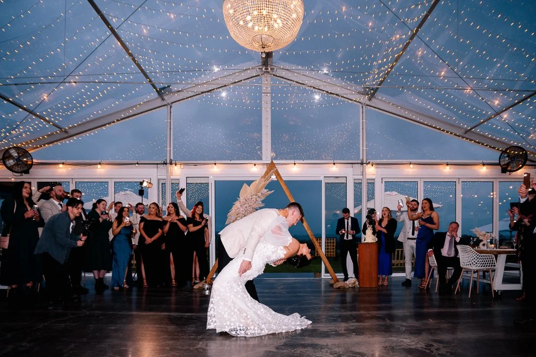 Bride and groom dancing at their wedding reception, surrounded by guests, under a decorated tent with string lights and a chandelier.