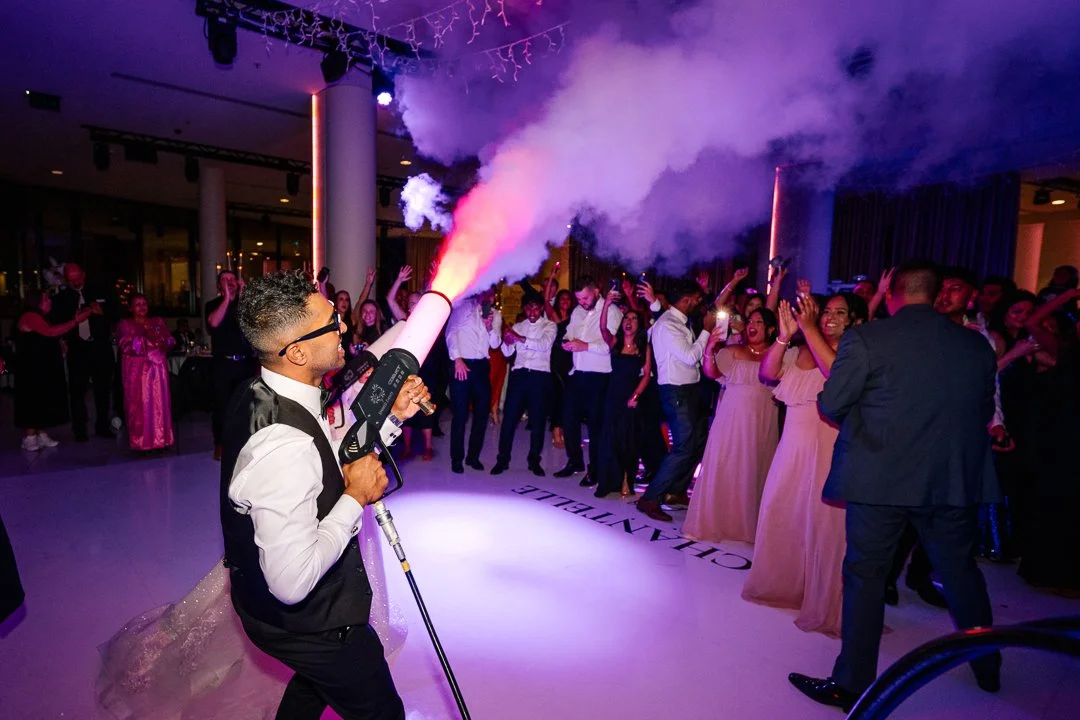 A DJ at a lively party holds a smoke grenade releasing a cloud of colorful smoke in front of a crowd of dancing people in formal attire.