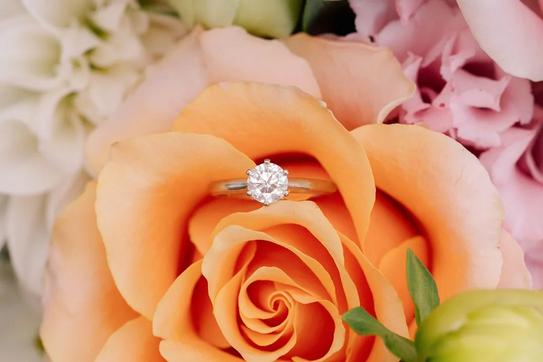 A sparkling diamond ring placed on an orange rose surrounded by pink and white flowers.